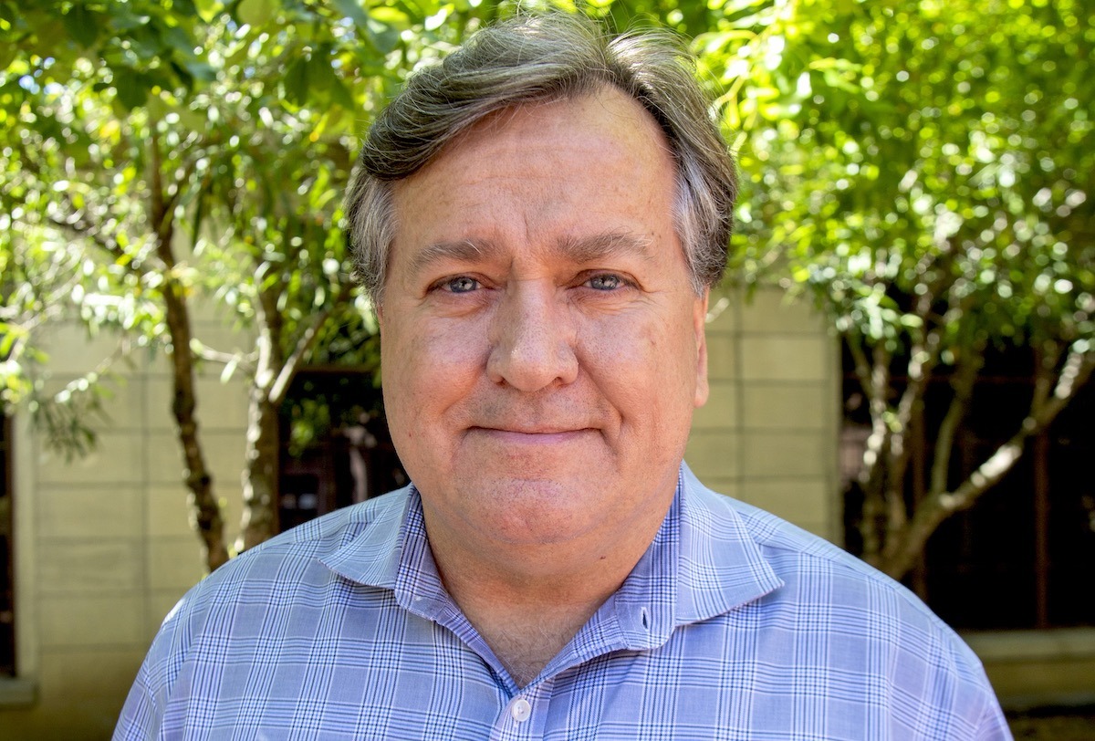 Portrait of a middle-aged person with graying hair, wearing a blue plaid shirt, standing outdoors in front of a background of green trees and a beige-colored building. The person, embodying the spirit of Berkeley Journalism, is looking directly at the camera with a neutral expression.