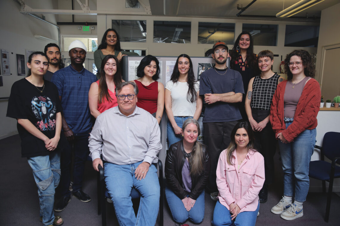 IRP group shot 1_ A group of 15 people posing for a photo in an office setting, some standing while others are seated. The group includes a mix of men and women of various ages, dressed casually. Dean Geeta Anand is among them. The background features office furnishings and wall decorations, perfect for the Quarterly Newsletter.