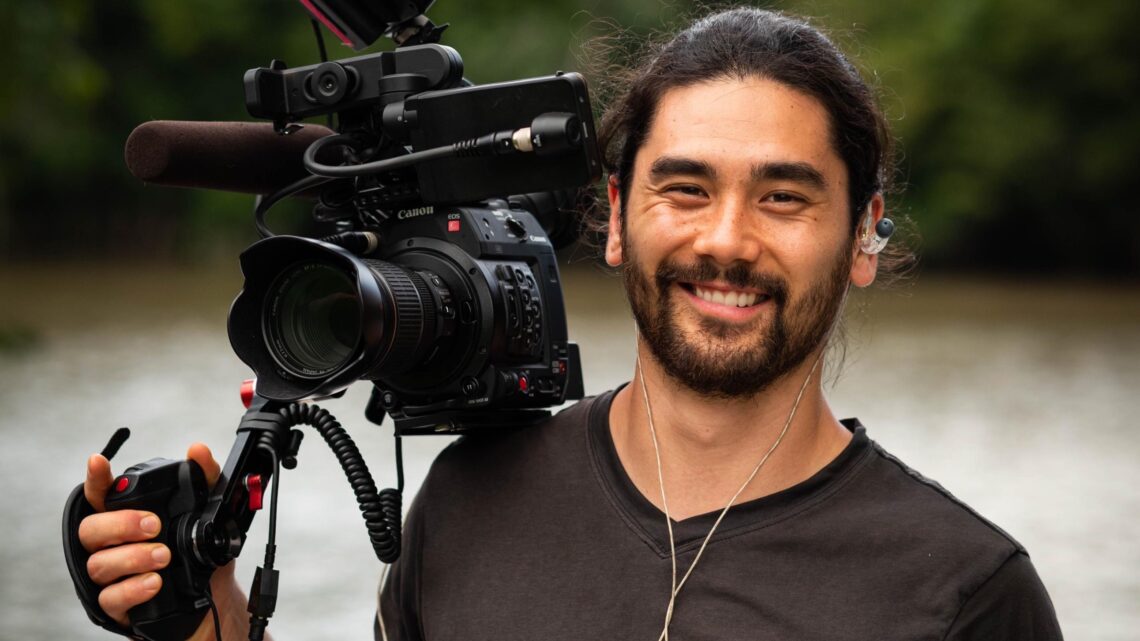A man smiles at the camera. He is holding a video camera that leans on his shoulder
